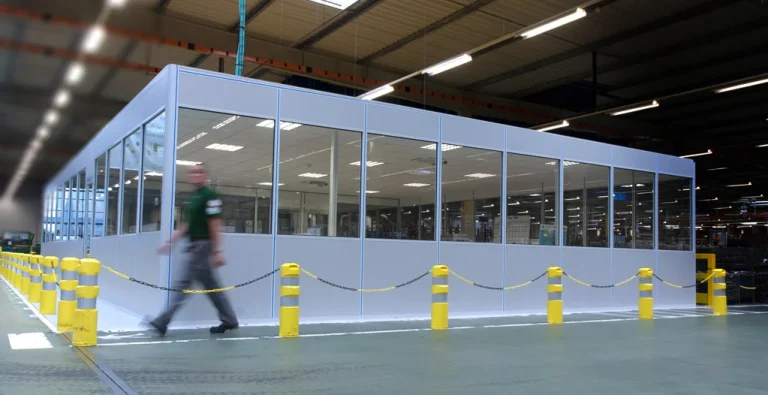 Longue cabine vitrée en kit dans un grand hall de production, protégée par des bornes et une chaîne de sécurité