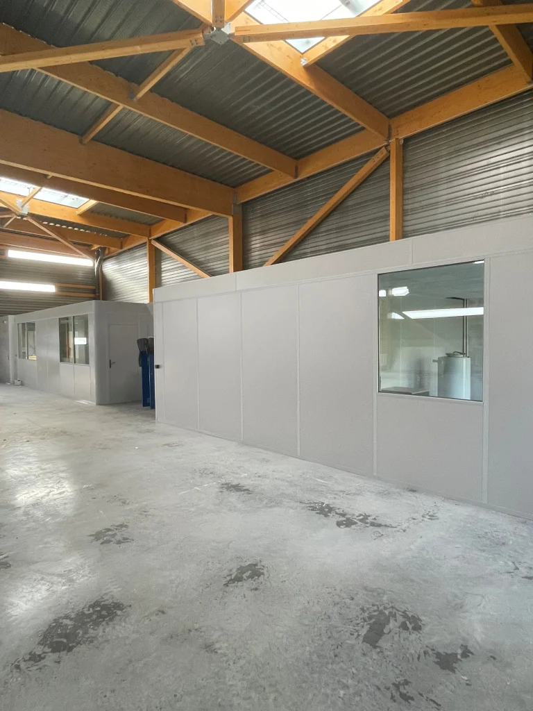 Cabine en kit à panneaux pleins et fenêtre vitrée, installée sous la charpente bois d’un entrepôt