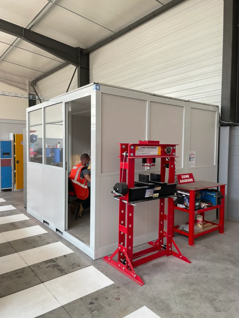Bureau d’atelier semi-vitré installé à côté d’une presse hydraulique et d’une zone de soudure