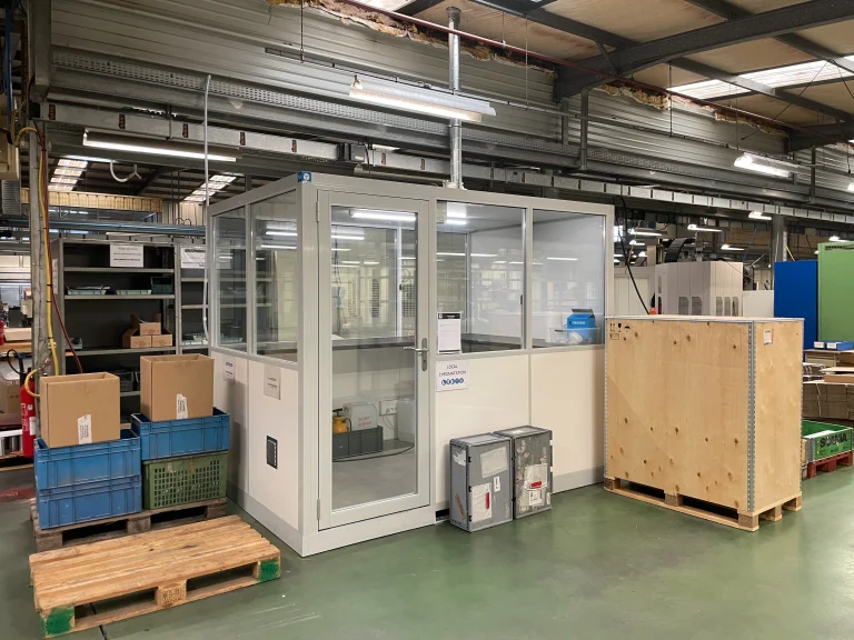 Poste vitré préfabriqué installé dans une zone de préparation logistique, entre palettes et caisses