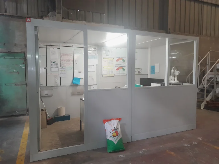 Cabine d’atelier en kit vitrée installée au pied d’un escalier métallique, dans un atelier au sol peint