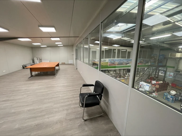 Plateau de bureaux à l’étage fermé par une cloison vitrée, avec vue sur l’entrepôt en contrebas